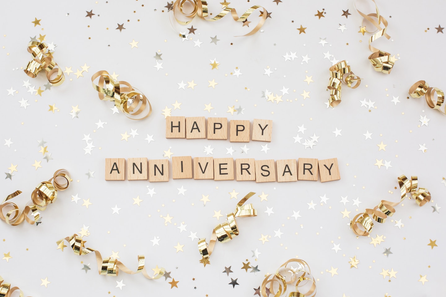 The words 'HAPPY ANNIVERSARY' in wooden letter tiles, surrounded by gold and silver coloured stars and gold curls, all set against a white background.