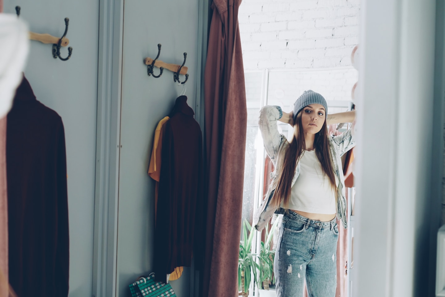 Mirror shot of young girl trying on clothes in fitting room. She is wearing denim jacket, jeans and hat, smoothing her hair, moving and looking in mirror to check image. Teenager shopping concept.